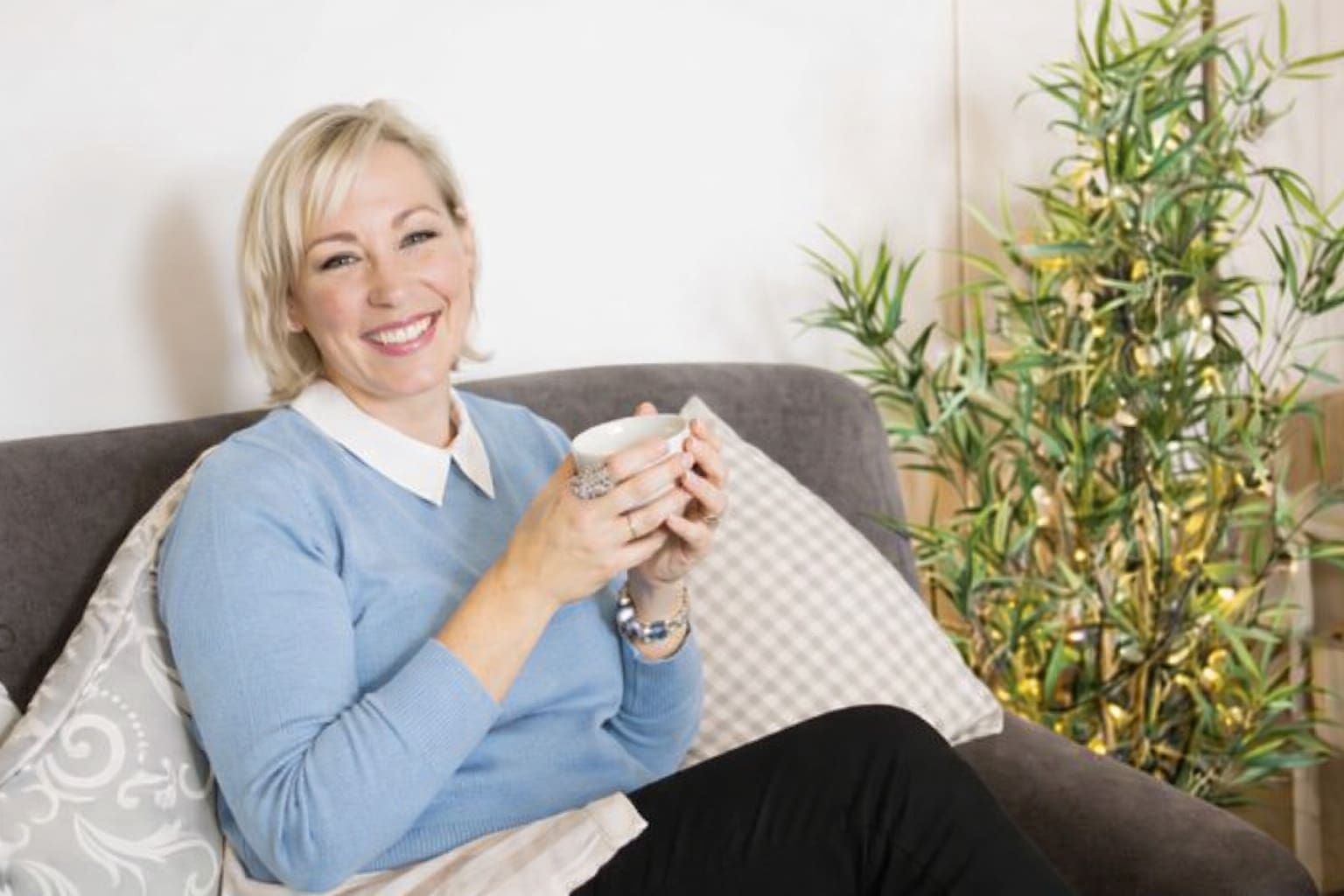 Woman with short blonde hair sits on a sofa, holding a mug and smiling, with a decorative plant and cushions in the background.