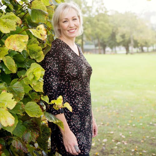 A woman with short blonde hair stands by leafy green bushes in a park, wearing a black patterned dress and smiling at the camera.